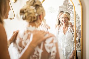 Bride laughing joyfully while admiring her lace wedding dress in an ornate gold mirror.
