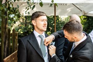Groomsman carefully pinning a boutonniere onto the groom's dark suit lapel outdoors.