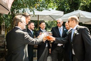 Six groomsmen in dark suits toasting with beer glasses outdoors under white umbrellas.