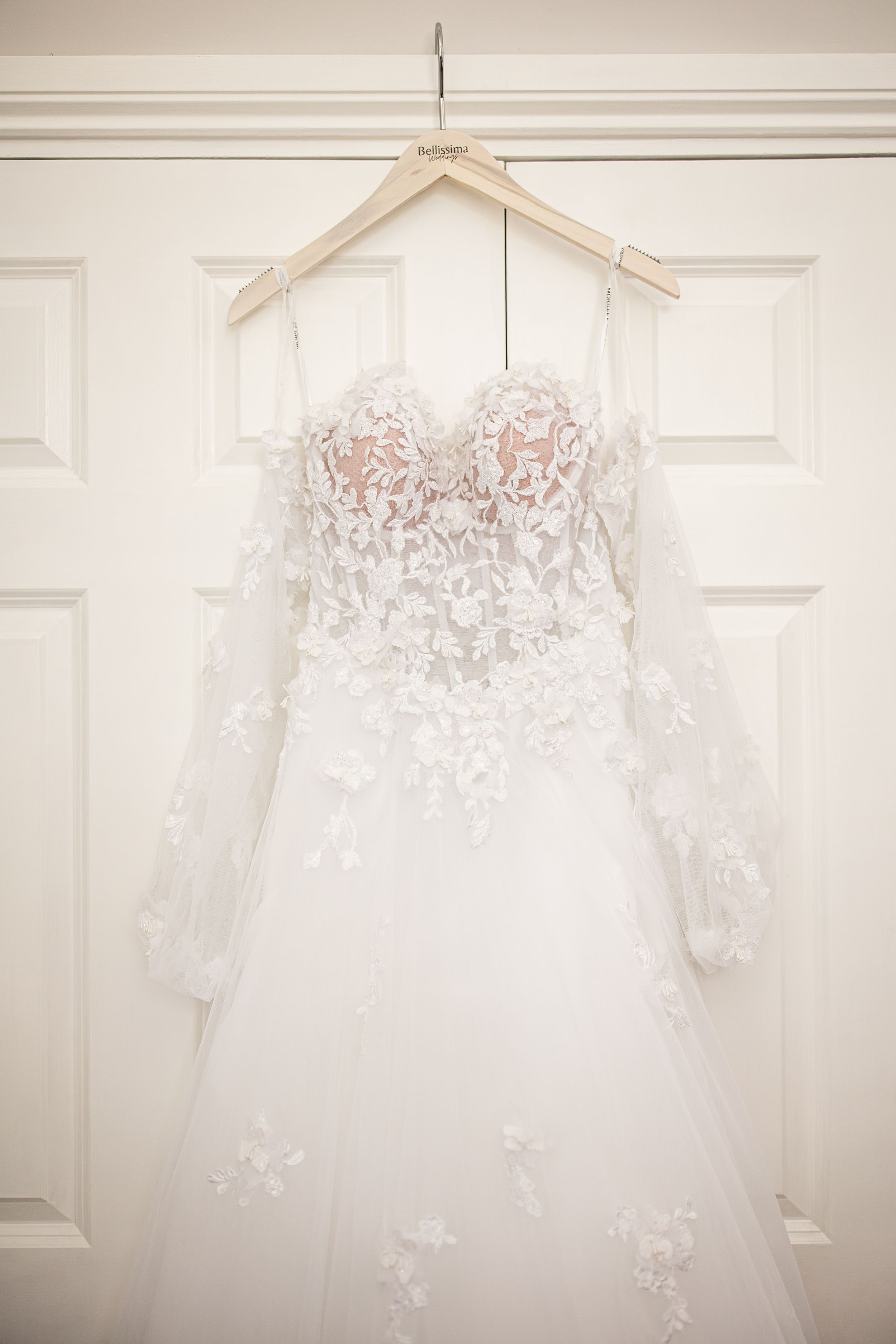 Elegant white lace wedding dress with floral applique and long sleeves hanging on a wooden hanger on a door.