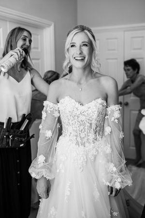 Radiant bride in off-shoulder lace wedding gown laughing while getting ready, black and white photo.