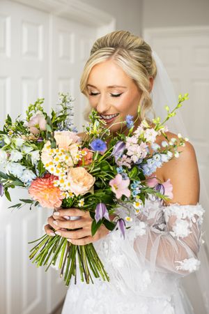 Smiling blonde bride in lace gown smelling a colorful wildflower wedding bouquet.