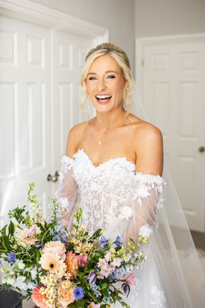 Joyful blonde bride in off-shoulder lace gown holding a colorful spring wedding bouquet.