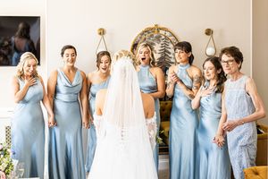 Bridesmaids in blue satin dresses reacting with awe to bride in white wedding gown and veil.