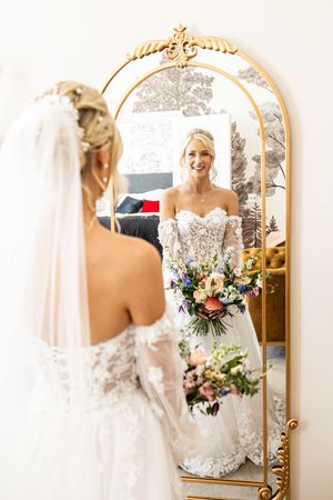 Bride in lace wedding gown and veil admiring her reflection in an ornate gold mirror.