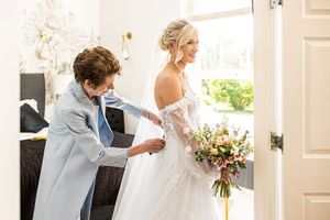 Mother helps bride with her lace off-shoulder wedding gown while holding a colorful bridal bouquet.