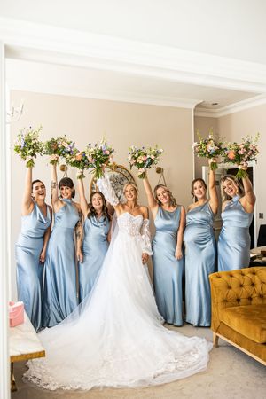 Bride and six bridesmaids in blue satin gowns joyfully raise colorful bouquets in a bright room.