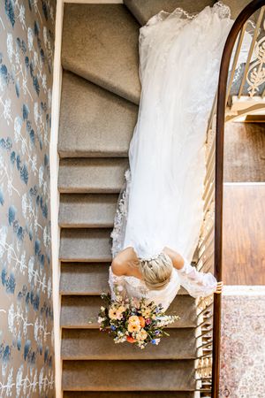Bride in lace wedding gown descending elegant spiral staircase holding colorful bouquet, viewed from above.