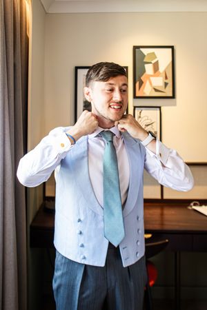 Smiling groom adjusting his mint green tie wearing a light blue waistcoat and white dress shirt with gold cufflinks.