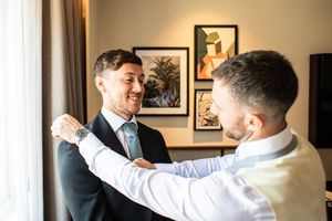 Smiling groom being helped into his dark suit jacket by a groomsman in a bright hotel room before the wedding.