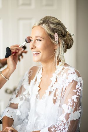 Blonde bride smiling while having makeup applied with a brush, wearing a white lace bridal robe during wedding prep.