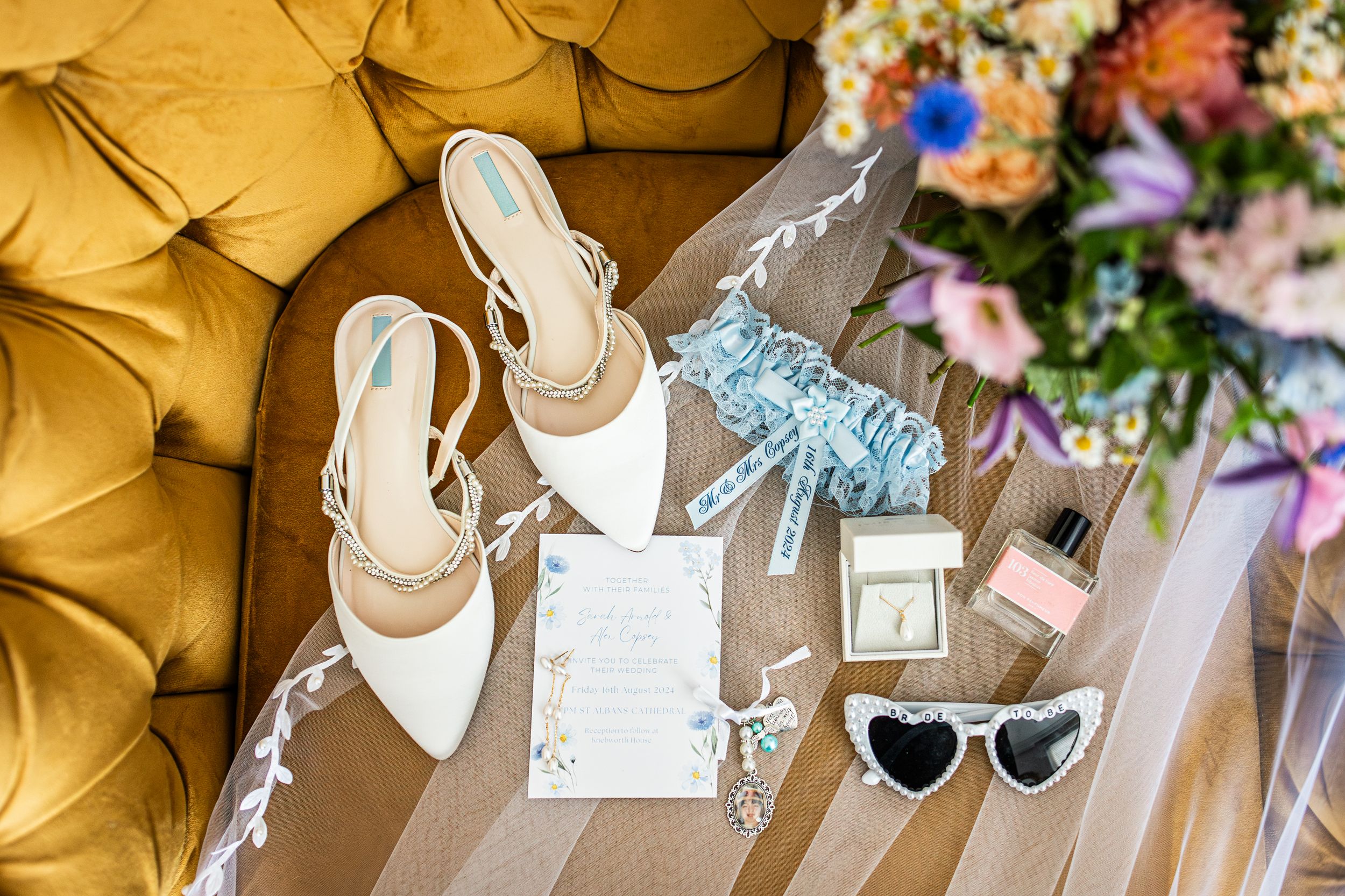 Bridal accessories flatlay including white heels, blue garter, heart sunglasses, and stationery on gold velvet sofa.