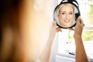 Beautiful blonde bride admiring her wedding makeup in a round handheld mirror on her wedding morning.