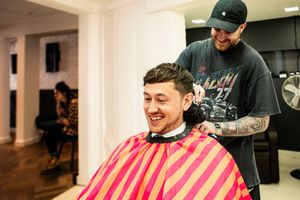 Groom laughing in a barber chair wearing a striped cape while getting a haircut before his wedding day.