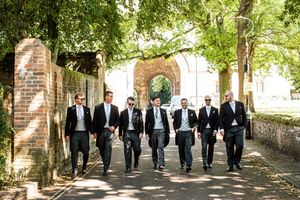 Groom and groomsmen in morning suits walking confidently along a tree-lined path near a brick archway.