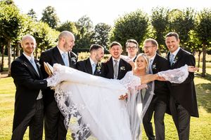 Groomsmen in black suits lifting bride in white wedding dress outdoors on sunny day.