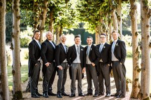 Group of groomsmen in black suits posing under trees at outdoor wedding venue.