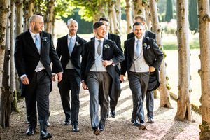 Groomsmen walking confidently in black suits along tree-lined path at wedding.