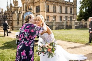 Bride in a white lace gown receiving a warm hug from a guest in a floral dress outside a historic manor.