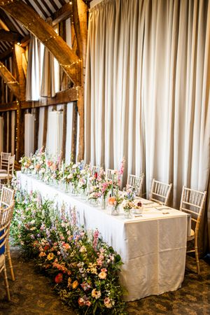 Long wedding head table decorated with colorful wildflower arrangements in rustic barn.