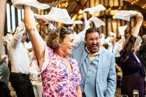 Wedding guests wave napkins in celebration at a rustic barn reception, with a woman in floral dress and man in blue suit laughing joyfully.