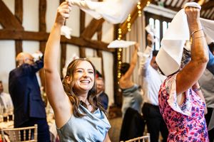 Smiling wedding guest in blue dress waves white napkin at elegant barn reception with fairy lights and other celebrating guests.
