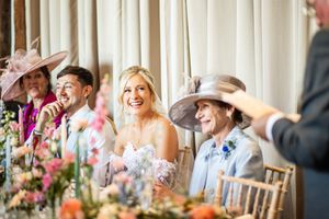 Radiant bride in strapless gown laughs with wedding party guests wearing decorative hats at floral-decorated head table.