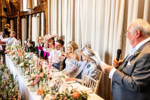 Wedding guests at floral head table react with delight as elderly man speaks into microphone at elegant reception.