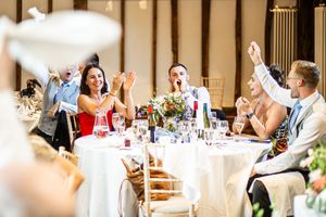 Excited wedding guests clap and cheer at round tables during lively reception in a decorated barn venue.
