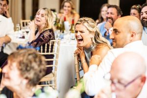 Wedding guest laughs heartily at reception table while surrounded by other amused guests in elegant venue.