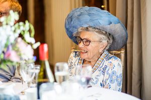 Elderly wedding guest smiles warmly wearing an elegant blue feathered hat at a floral-decorated reception table.