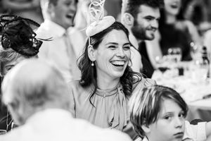 Black and white photo of laughing wedding guest wearing fascinator hat at elegant reception with guests in background.