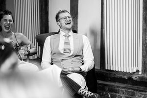 Black and white photo of groom laughing heartily in waistcoat and tie during wedding reception speeches.