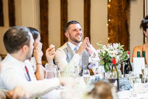 Smiling wedding guest in grey suit clapping during speeches at an elegant wedding reception.