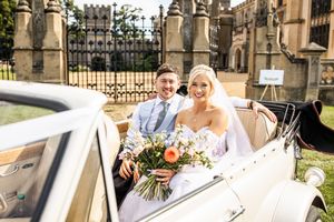 Smiling newlyweds sit together in a vintage white convertible car outside a historic cathedral.