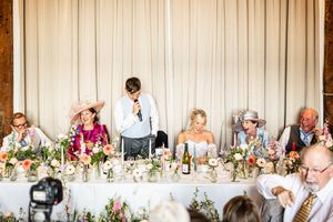 Wedding party seated at top table during speeches at a floral-decorated barn wedding reception.
