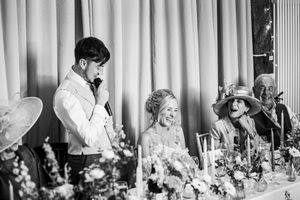 Black and white photo of groom giving speech while bride laughs at wedding top table.