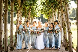 Bride and bridesmaids in blue satin dresses walking through a tree-lined avenue holding colorful bouquets.