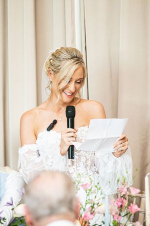 Blonde bride smiling while giving speech holding microphone and notes at wedding reception.