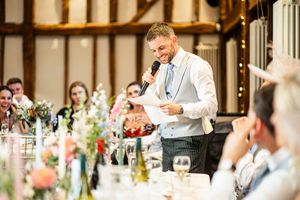 Groom in grey suit delivering wedding speech at floral-decorated barn reception venue.
