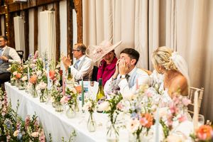 Wedding party reacting with laughter at top table decorated with pink floral arrangements.
