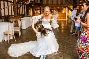 Bride in lace gown with tiara dancing and twirling young flower girl in blue dress inside rustic barn venue.