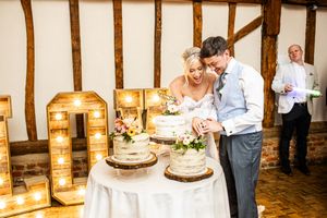 Bride and groom cutting a rustic naked wedding cake beside illuminated letter lights in a barn venue.