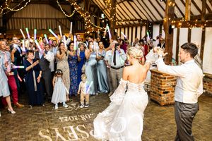 Bride and groom enter reception as guests cheer with glow sticks in a decorated barn wedding venue.