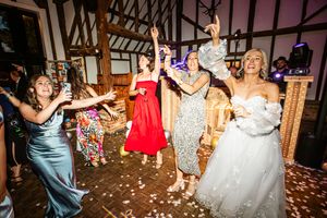 Bride and bridesmaids dancing joyfully at wedding reception in rustic barn venue with confetti.