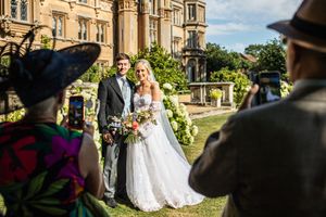 Bride and groom posing for photos in front of a grand manor house while guests capture the moment.