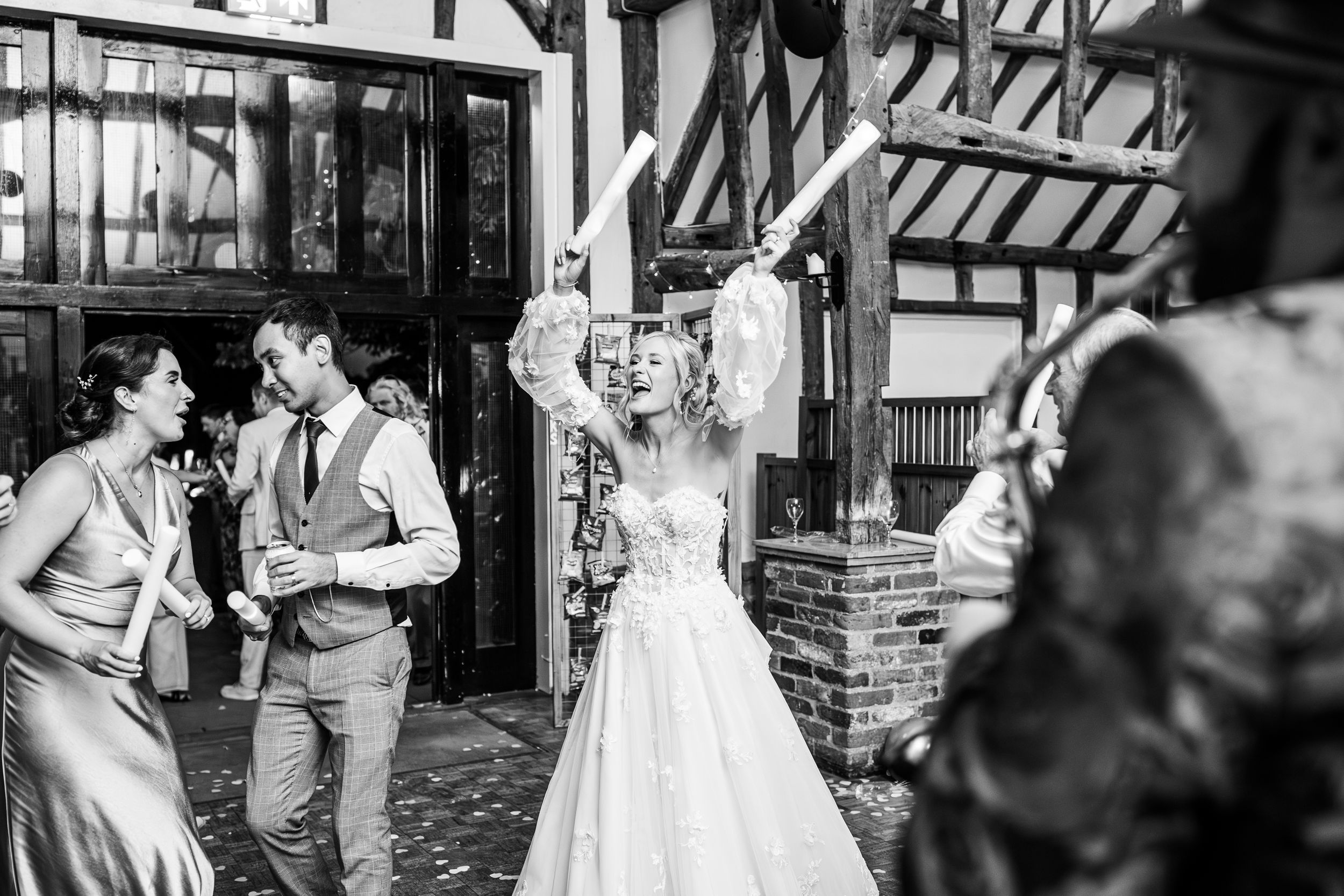 Bride in white gown celebrating with guests outside Tudor-style wedding venue in black and white.
