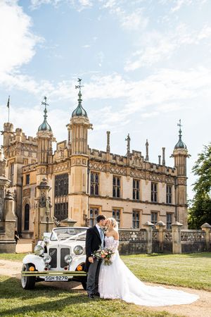 Bride and groom kissing beside a vintage white car in front of a grand English manor house on their wedding day.