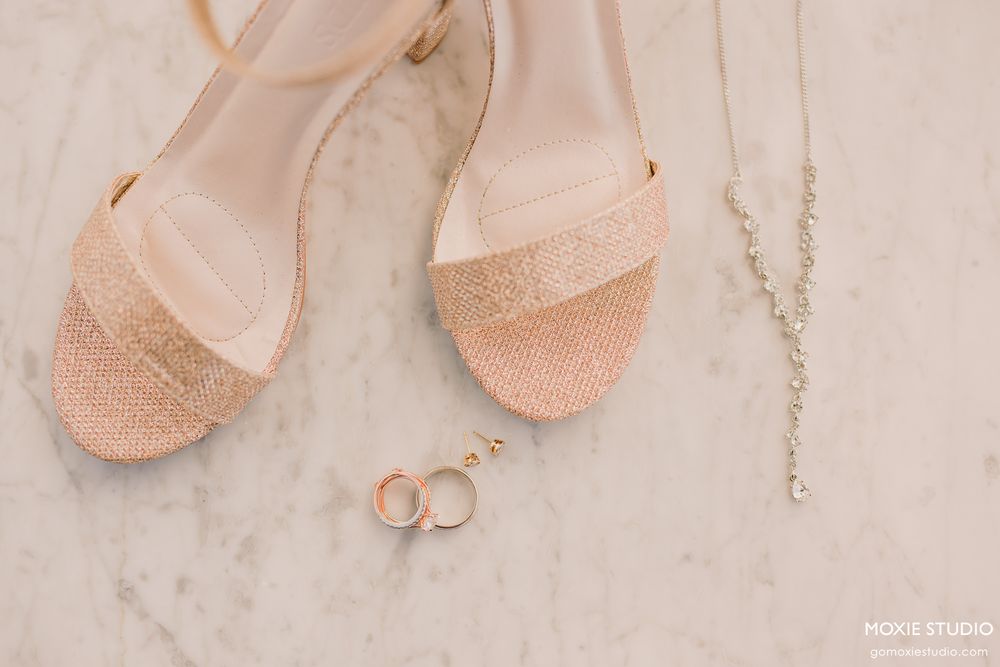 Wedding accessories including sparkly blush sandals, wedding rings and a delicate necklace arranged on marble surface.