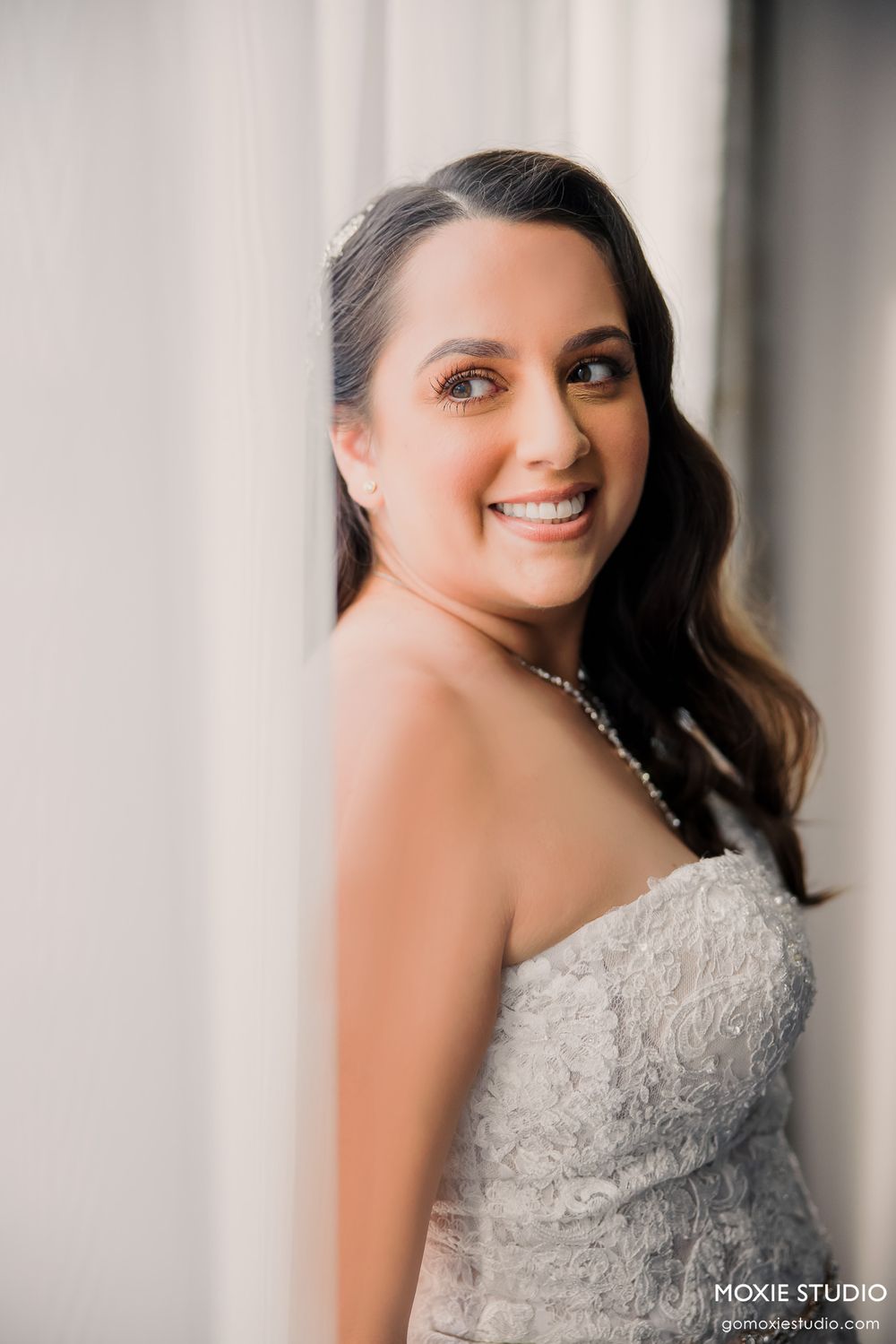 Elegant bride wearing a detailed lace strapless wedding gown poses by a window.